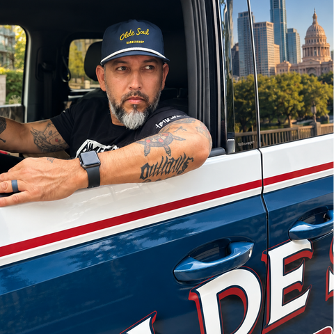 A man with a beard and tattoos leans out of a blue and white vehicle window, wearing a black t-shirt and a navy blue cap with 'Olde Soul barbershop on it. The vehicle has 'OLDE SOUL BARBERSHOP AUSTIN TX' printed on the side.